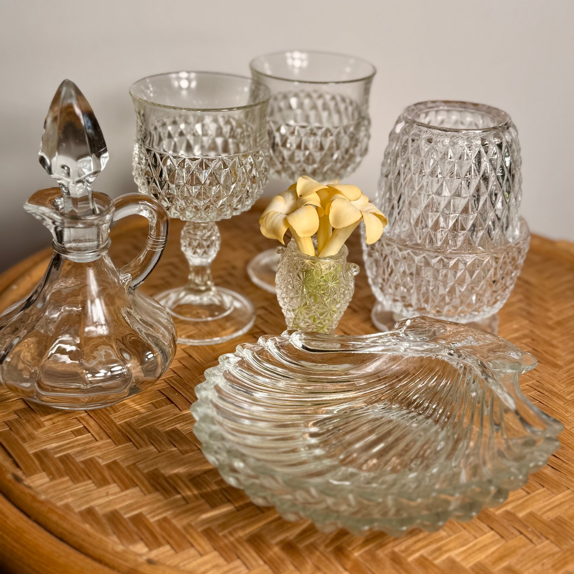 A softly styled flatlay featuring the full Quiet Luxe Collection: a curated mix of clear vintage glass captured in golden natural light atop a woven lauhala table. The grouping includes a faceted cruet with stopper, a matching pair of Indiana Glass diamond point goblets, a sparkling fairy lamp, a pair of shell-shaped Fitz & Floyd serving dishes, and a tiny Daisy & Button bud vase cradling real plumeria. The textures, curves, and clarity reflect timeless